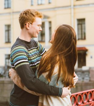 Girl with long thick dark hear embracing redhead boy in the blue t-shirt on a bridge, young couple. Concept of teenage love and first kiss, sincere feelings of man and woman, city, waterfront. Vertical