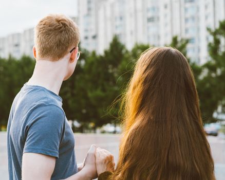 Girl with long thick dark hair holding hands redhead boy in blue t-shirt on bridge, teen love at evening. Boy looks tenderly at girl, young couple. Concept of teenage love and first kiss, sincere feelings of man and woman, city, waterfront. Close up