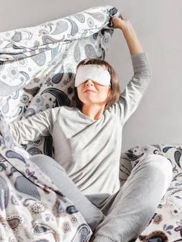 Young woman in grey pajama and sleeping mask on face. She is just woke up and sit in bed. Early morning in cozy home.