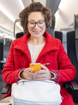 Smiling woman in red duffle coat texting on smartphone in suburban train. Travel by land vehicle.