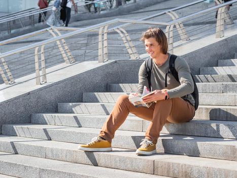Smiling man sits on stone staircase in park and reads travel guide. Solo-travelling around city. Urban tourism. Modern architectural landmarks.