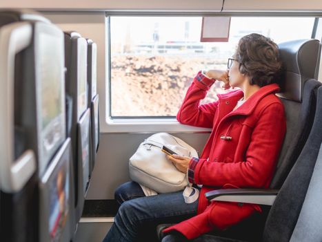 Smiling woman in red duffle coat with on smartphone sits near window in suburban train. Travel by land vehicle.