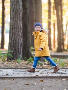 Little boy in yellow jacket runs on walking path at park. Autumn sunny day. Leisure activity outdoors. Soft focus.