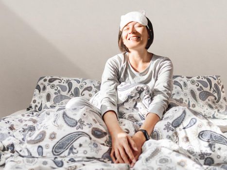 Smiling woman in grey pajama and sleeping mask. She is just woke up and sit in bed. Early morning in cozy home.