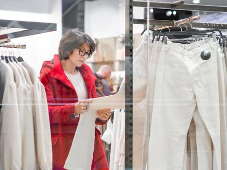 Woman chooses trousers at clothing store. Casual trousers hanging on hangers. View through transparent shop window. Shopping at mall. Basic clothes for everyday wear. Modern urban fashion.