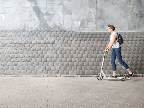 Woman rides kick scooter. Female in jeans and sneakers moves fast on grey tiled wall background. Healthy lifestyle. Eco-friendly urban transport.