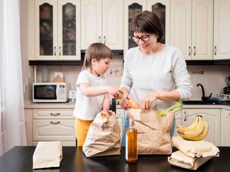 Woman and toddler boy sorts out purchases in the kitchen. Mother and son with grocery delivery in paper bags. Subscription service from grocery store.