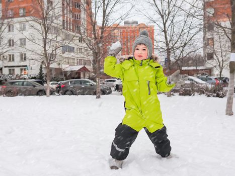 Laughing boy in green jumpsuit plays snowball on snowy yard among trees. Leisure activity for children in winter.
