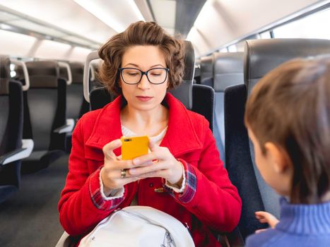 Curly woman in red duffle coat is texting on smartphone in suburban train. Woman sits with her son. Travel by land vehicle.