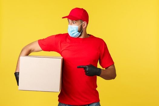Express delivery during pandemic, covid-19, safe shipping, online shopping concept. Startled and shocked courier in red uniform, medical mask and gloves, pointing left, holding box package