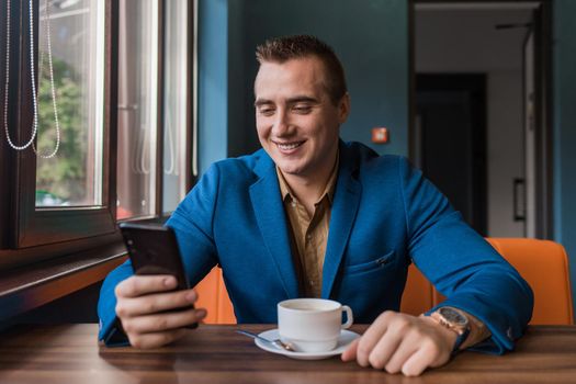 A young smiling positive handsome guy businessman of European appearance portrait, uses a smartphone or mobile phone sitting in a cafe at a table on a coffee break