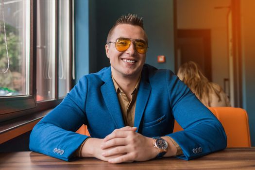 Cute positive smiling businessman of Caucasian appearance stylish portrait in sunglasses, jacket and shirt, sitting at a table in a cafe idly by