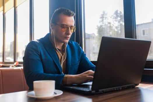 A business man stylish serious businessman in an attractive European-looking suit works in a laptop, listens to music with headphones and drinks coffee sitting at a table in a cafe by the window