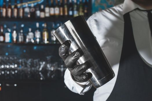 The hand of a professional bartender in a black latex glove with a bar instrument for mixing and preparing alcoholic cocktails with a shaker