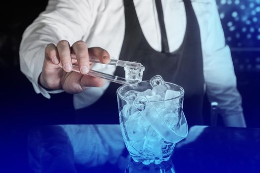 The hand of a professional bartender holds plastic ice cube tongs next to a cold ice dish