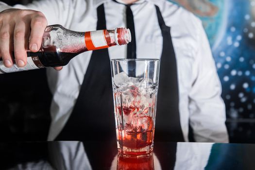The hand of a professional bartender pours red syrup into a glass of ice cubes. The process of preparing an alcoholic cocktail