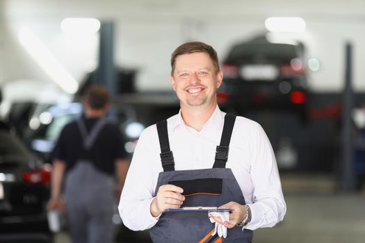 Smiling maintenance center worker in uniform, man fill in papers on car checkup