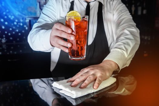 A professional bartender holds in his hand a red alcoholic cocktail, chilled drink in glass decorated with a slice of sliced lemon and wipes the bar counter with white cloth for serving
