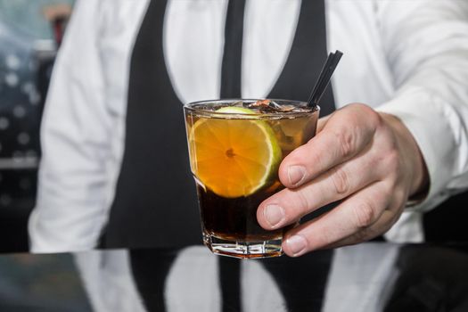 The hand of a professional bartender serves an alcoholic beverage cocktail whiskey with cola and a piece of lime on the bar counter, close-up