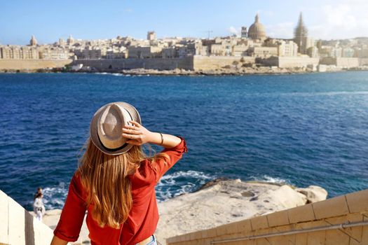 Beautiful young woman with hat descends stairs in Malta towards waterfront looking at panoramic view of Valletta, Malta