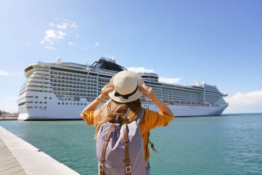 Young female backpacker holding straw hat standing in front of big cruise liner