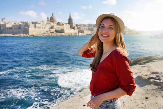 Portrait of young woman stands on waterfront looking at camera with Valletta city on the background, Malta Island