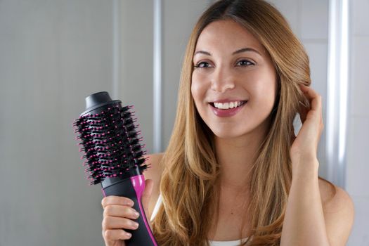 Pleased girl holds round brush hair dryer to style hair in her bathroom at home. Young woman showing salon one-step hair dryer and volumizer.