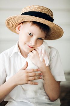 Front view of boy wearing white shirt and hay hat keeping cute white bunny and smiling. Young male hugging rabbit and posing while resting in village. Concept of childhood, animals and love.