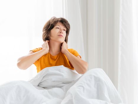 Sleepy woman in yellow pajama is massaging her neck. Waking up early in morning. Woman gets enough sleep.