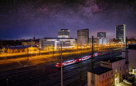 High speed train in motion and Milky Way at starry night. Industrial landscape with sky and stars over blurred modern passenger train and railroad.