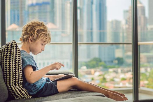 Boy uses a tablet at home on the couch in the background of a window with skyscrapers. Modern children in the megalopolis use a tablet concept