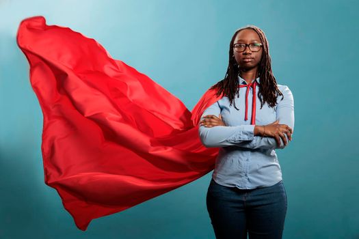 Confident and brave young superhero woman standing with arms crossed while wearing red mighty cape