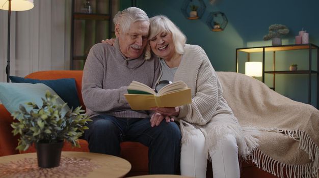 Senior grandparents couple relaxing, reading book, talking enjoying leisure hobbies at night home