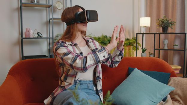 Young Caucasian girl wearing VR headset resting at home sitting on sofa in living room alone using futuristic technology making gestures with hands in air, play 3D video game. Virtual reality glasses