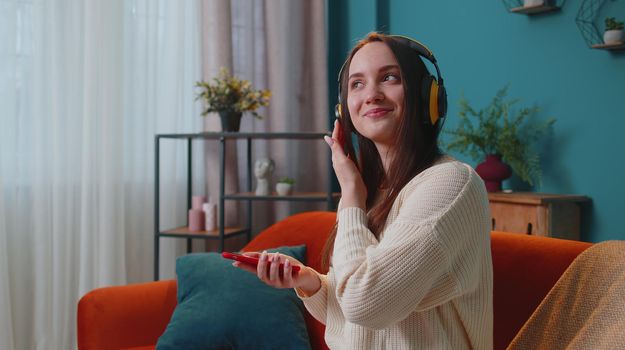 Pretty Caucasian young woman in headphones listening music dancing, singing in living room at home