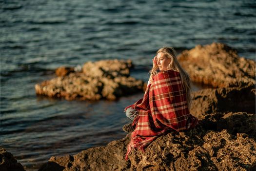 Attractive blonde Caucasian woman enjoying time on the beach at sunset, sitting in a blanket and looking to the side, with the sunset sky and sea in the background. Beach vacation.