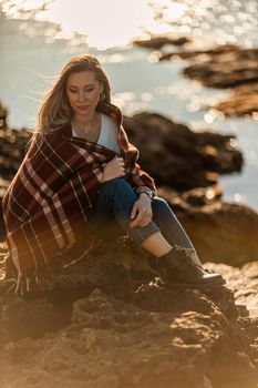 Attractive blonde Caucasian woman enjoying time on the beach at sunset, sitting in a blanket and looking to the side, with the sunset sky and sea in the background. Beach vacation.