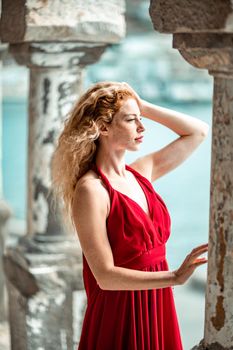 Outdoor portrait of a young beautiful natural redhead girl with freckles, long curly hair, in a red dress, posing against the background of the sea.