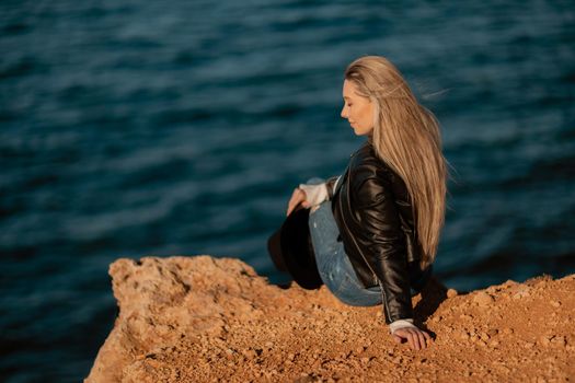 A blonde girl in a stylish black leather jacket is sitting with her back to the seashore.