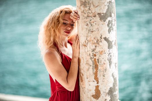 Outdoor portrait of a young beautiful natural redhead girl with freckles, long curly hair, in a red dress, posing against the background of the sea.