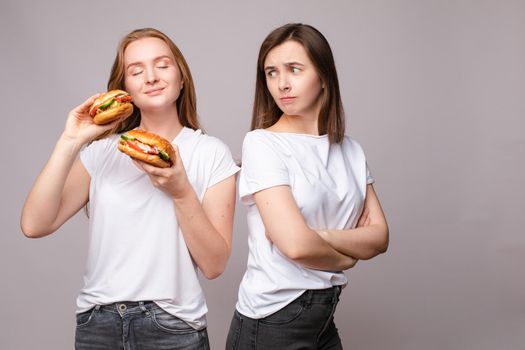Two frowning young beautiful woman holding appetizing harmful burger looking at camera