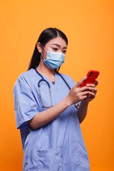 Healthcare clinic nurse wearing virus protection facemask and medical instrument while using smartphone for messages. Medical caregiver wearing stethoscope and protective mask while having phone.
