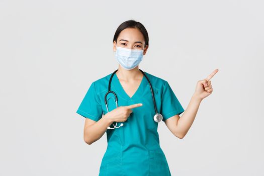 Covid-19, coronavirus disease, healthcare workers concept. Pleasant asian female doctor, medical employee in scrubs and mask, pointing fingers upper right corner, showing advertisement