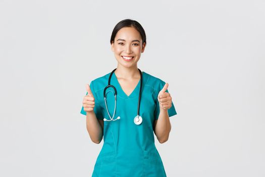 Covid-19, healthcare workers, pandemic concept. Professional confident smiling doctor, female physician in scrubs showing thumbs-up with assured expression, ensure all good, perfect or excellent