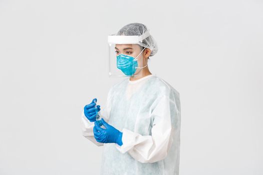 Covid-19, coronavirus disease, healthcare workers concept. Side view of serious young asian female doctor, physician in personal protective equipment insert syringe into ampoule with vaccine