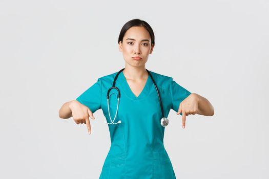 Covid-19, healthcare workers, pandemic concept. Upset and disappointed pouting asian female therapist, nurse in scrubs pointing fingers down, sulking sad, showing something with regret