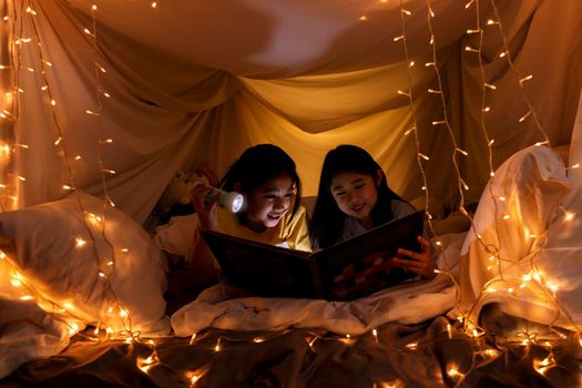 Family concept. elder sister and sister reading book with flashlight together in children tent before bedtime. Happy elder sister read story book to her sister in bed sheet tent
