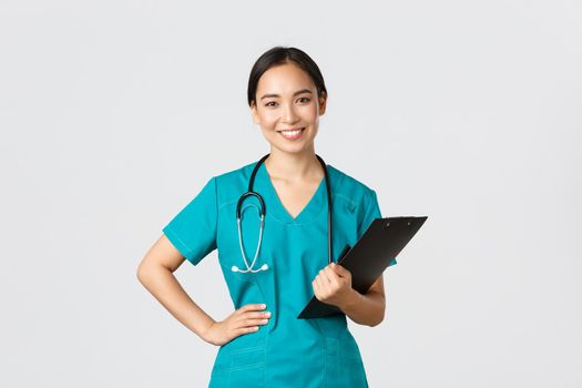 Healthcare workers, preventing virus, quarantine campaign concept. Smiling pleasant asian female physician, doctor during examination wearing scrubs and holding clipboard, white background