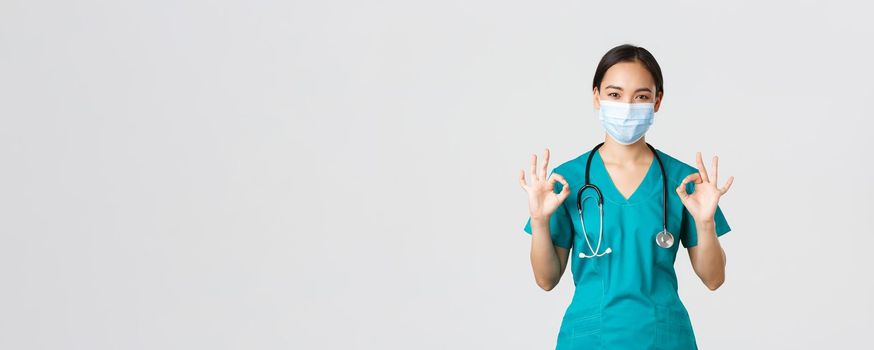 Covid-19, coronavirus disease, healthcare workers concept. Young professional female doctor, asian intern in scrubs and medical mask showing okay gesture, guarantee quality