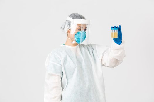 Covid-19, coronavirus disease, healthcare workers concept. Side view of serious-looking tech lab employee, researcher in personal protective equipment examine urine analyzes, white background